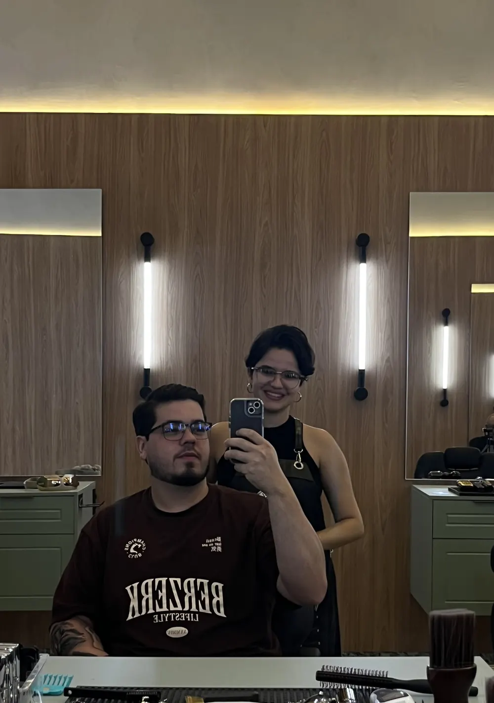 Cliente na cadeira com a barbeira ao lado, refletidos no espelho iluminado