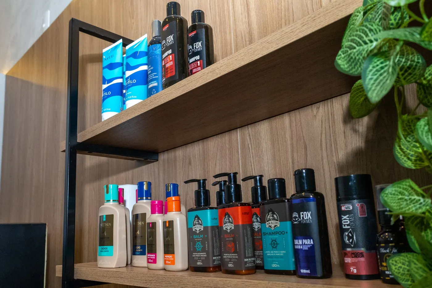 Prateleira de madeira com frascos de produtos para cabelo e barba ao lado de uma planta